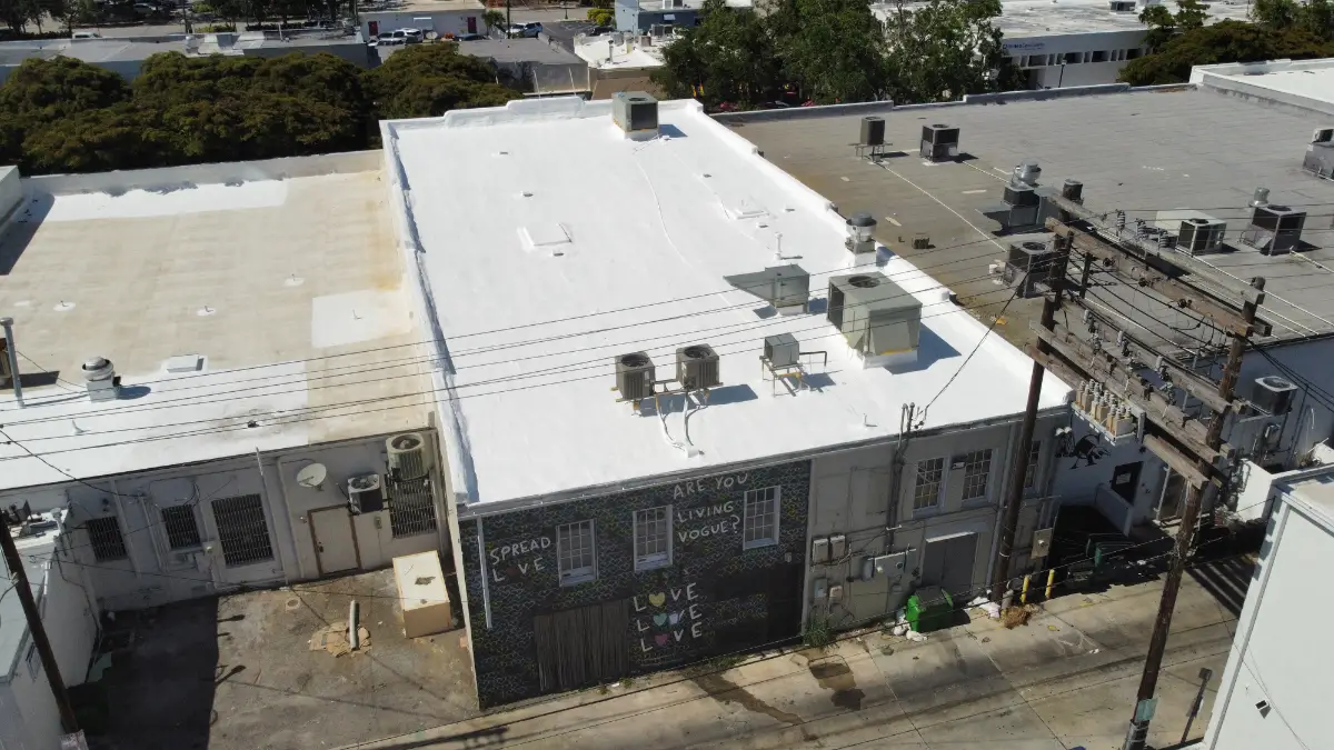 white-commercial-flat-roof-restoration Aerial view of a commercial flat roof with a bright white restored roof surface, rooftop HVAC units, neighboring buildings, and utility lines